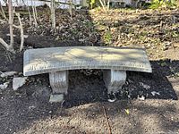 Concrete garden bench placed outside on bare soil with leaf litter in the background. Shows curved seating surface and carved decorative edging.