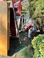 Photo shows a green metal tine garden rake with a wooden handle leaning against a wooden table outdoors, surrounded by various garden and household items.