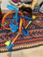 Hot Wheels car track set assembled on a patterned rug with blue, orange, and yellow components forming a loop and launcher mechanism.