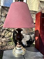 Lamp shown with maroon conical fabric shade and wooden turned base with metal bottom, photographed outdoors with natural light.