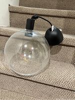 Clear glass globe pendant lamp resting on carpeted stairs. Shows transparent spherical glass shade, light bulb inside, black canopy and cord.