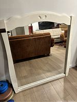 Frontal image showing the white wood framed rectangular mirror standing on the floor against a wall with some furniture reflected in the glass.