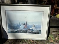 Framed painting showing three children sitting on large rocks by water, one child pointing up to the sky with a scenic backdrop of trees and mountains.