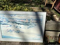 Framed painting showing a panoramic beach scene with multiple beach chairs and coastal buildings under a cloudy sky.