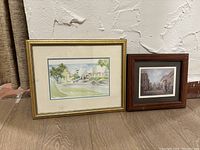 Two framed pictures on the floor against the wall. Left one is a watercolor nature and building scene with a gold frame, right one is a dark wood framed street view monochrome print.