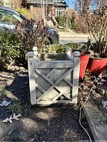 White wooden plant pot with square top, crisscross design and corner finials, sitting outdoors.