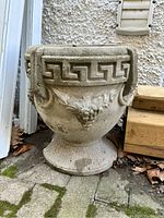 Concrete decorative planter with Greek key design around the rim and grapevine relief motif on the main body. Positioned on stone or concrete pavement with some dried leaves around.