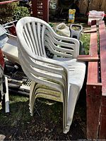 Four white plastic outdoor chairs stacked together outside on the grass with various outdoor items nearby. Chairs are dirty and need cleaning.