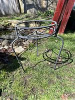 Photo showing a black rod iron bird bath holder stand placed on grass with a wooden fence in the background. The stand features a circular top ring designed to hold a bird bath with decorative curved ironwork beneath.