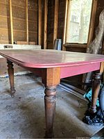 Side view of painted oak dining table showing red top and natural finish turned legs on concrete floor and unfinished wooden walls background.
