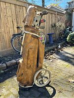 Full side view of brown vintage leather golf bag mounted on two-wheel pushcart. Contains multiple golf clubs with wooden and metal heads. Bag shows wear consistent with age and use. Photo taken to show entire lot composition and condition.