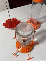 Two Perky-Pet red hummingbird feeders on a white surface, one with clear glass nectar container and the other just the red plastic base.