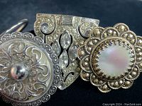 Close-up photo showing three vintage ornate dress shoe clips: one round silver-toned with filigree and dome, one rectangular with crystals, and one pair round with mother-of-pearl center and scalloped rhinestone frame.