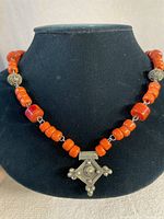 Front view of the necklace displayed on a black bust showing red-orange beads, brass beads, and square brass pendant