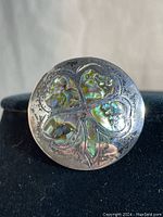 Front view of round sterling silver pendant brooch with four-leaf clover inlayed with abalone shell, showing iridescent colors and patina variegations on the metal.