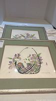 Two framed watercolor artworks showing a wire basket with lavender flowers, framed with glass and a green mat.