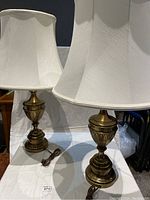 Full view of two matching brass base table lamps with off-white fabric shades, cords coiled beside them.