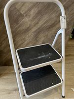 Metal folding two-step stool angled front view showing top and bottom steps with black ribbed rubber surface and white metal frame