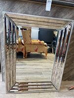 Full view of the rectangular metal frame wall mirror showing the stepped reflective metal border and the reflective glass center reflecting an armchair and floor.