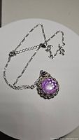 Front angled view showing the large round purple amethyst pendant with decorative silver setting and chain scattered around pendant.