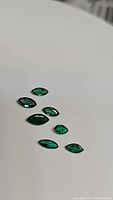 Seven marquis cut emerald gemstones with vivid deep green color laid out on a white surface.