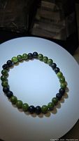 Top-down view of the natural green jade and black lava stone bead bracelet placed on a white circular surface under lighting that highlights bead textures.