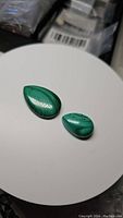 Two pear-shaped cabochon natural malachite gemstones on a white round surface showing their characteristic banded green coloration and smooth polished finish.