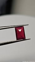 Close-up view of a rectangular baguette cut natural ruby held with tweezers showing its deep red color and faceted surface.