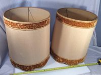 Two large beige fabric lamp shades with brown velvet and gold braided trim, standing side by side on a gray surface.