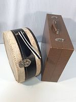 Two vintage carrying cases side by side, one rectangular wooden case with metal clasps and handle, and one round black and white case with wrist strap