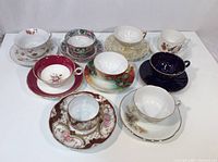 Top view showing 9 teacups and saucers in assorted floral and decorative patterns, colors include red, cream, dark blue, brown and multicolor accents.