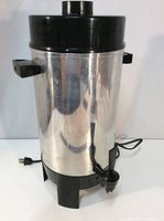 Front view of electric coffee percolator showing metal body, black plastic top with knob, two side handles, spout at base, and power cord.
