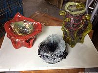 Top-down view of three colorful art glass sculptures on table: red bowl, grey/black bowl, and green vase.