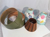 Photo showing all four lamp shades together: green fabric shade, brown wooden slat shade, and two small cartoon-themed shades with pastel colors and checkered patterns.