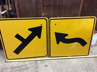 Two yellow metal street signs side by side on floor, showing straight/right and left curves