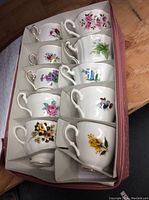 Photo showing 10 colorful vintage porcelain teacups in cardboard box, each with different floral patterns and decorative handles.