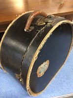 Side profile of vintage black round hatbox showing leather handle and lock mechanism with some wear on edges.