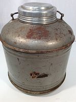 Full view of the vintage metal Thermos jug showing its cylindrical shape, domed lid, and metal handles with rust and wear.