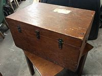 Closed vintage wooden tool/storage box showing the rectangular shape, dark finish, wood grain, and metal clasps on the front. The surface has wear and scratch marks.