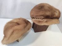 Two brown women's fur winter hats, one styled with a chain embellishment and rounded shape, the other smaller rounded fur hat.