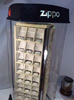 Front view of the Zippo lighter display case showing the clear plastic enclosure, white internal rotating panels, black base and top with Zippo logo and lock.