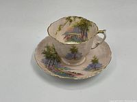 Tea cup and saucer set showing scenic landscape with trees and bridge, gold trim on handle and edges.