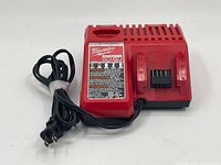 Front side of red Milwaukee battery charger with power cord coiled beside it, showing label and prongs for battery attachment.