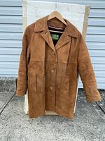 Front view of the brown suede jacket hanging on a wooden hanger outdoors, showing button closure and collar.