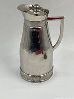 Overall view of vintage metal Thermos pitcher with hinged lid and handle on white background, showing reflective silver-tone finish and shape.