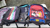 Five backpacks displayed on a table, showing varied designs including two PlayStation backpacks and one colorful striped backpack.