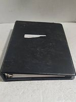 Closed black three-ring binder with a partially torn white label on front cover.
