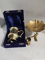 Brass pitcher, flower-shaped pedestal stand, and small cup arranged in and near blue velvet lined case