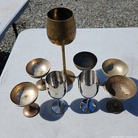 Top and angle view of 8 metal goblets and cups made of brass and copper with varying heights and finishes on white table.