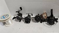 Five fishing reels lined up horizontally with a spool of fishing line on the left side on a white background.